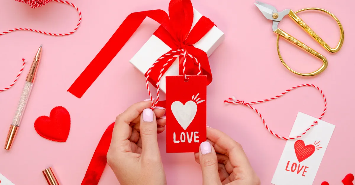 Hands holding a red ‘Love’ gift tag attached to a white present with red ribbon, surrounded by hearts, scissors, and wrapping accessories on a pink background.