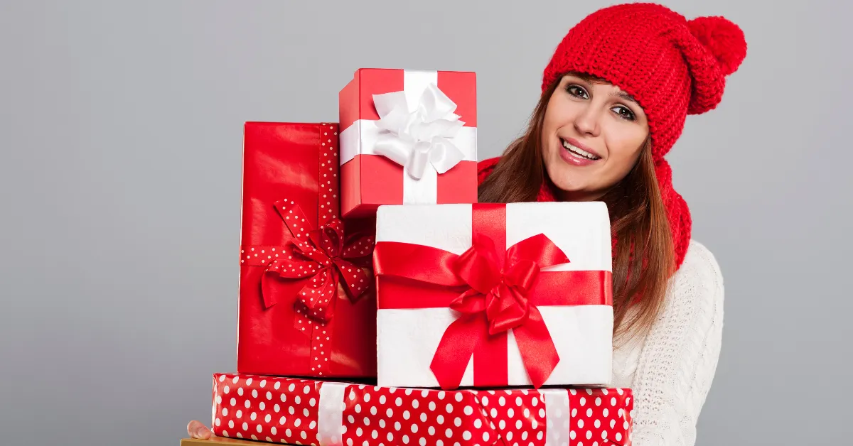 A smiling woman wearing a red winter hat holding several red and white wrapped gift boxes with ribbons.