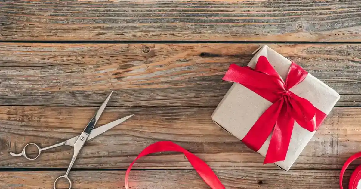 A gift box wrapped with a red ribbon on a wooden table, with scissors and a loose red ribbon nearby.