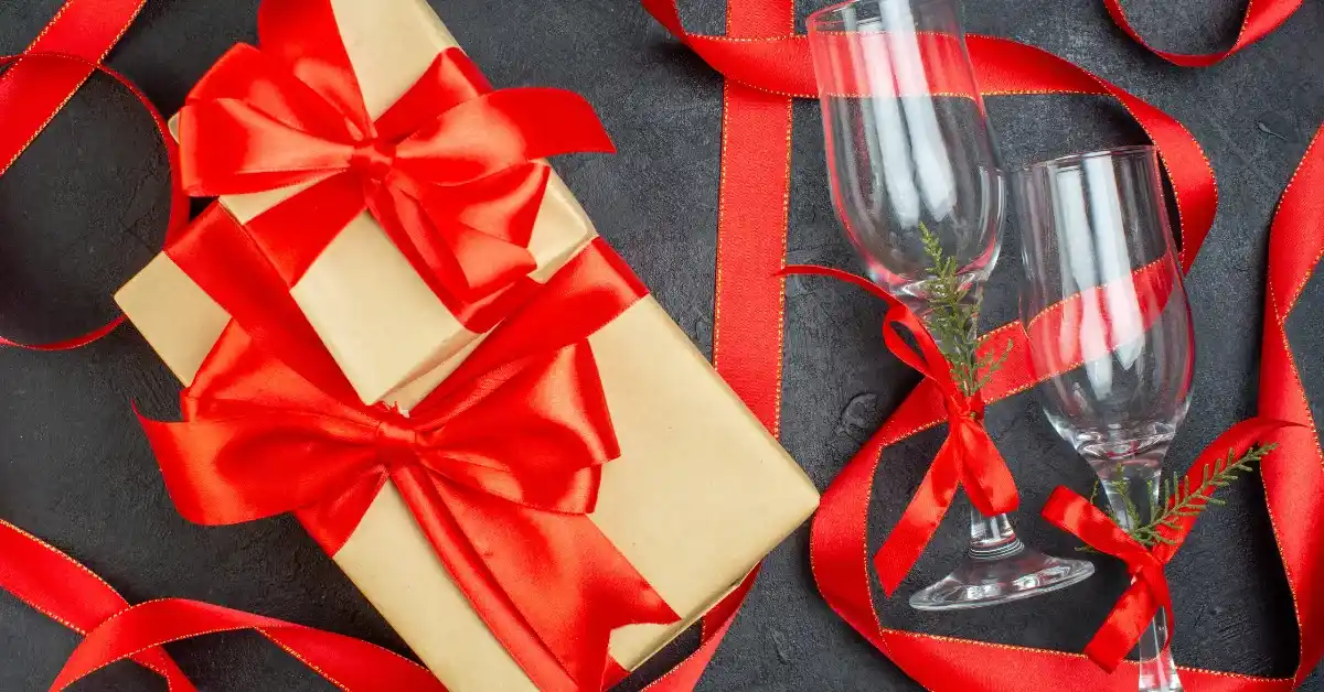 A gift box tied with a red ribbon and two champagne glasses, with red ribbons and small green sprigs on a dark background.