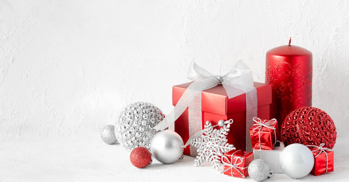 Red gift box with a silver ribbon surrounded by Christmas ornaments, including red and silver baubles, small wrapped presents, a decorative snowflake, and a red candle on a white background.