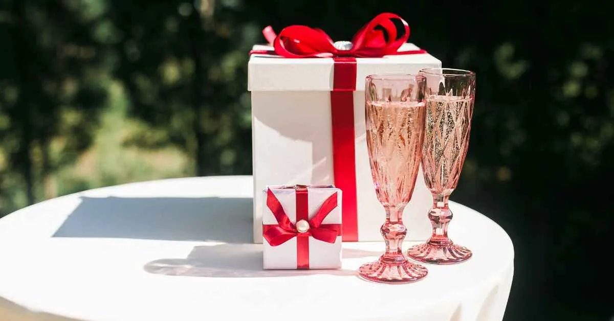 White gift boxes with red ribbon bows placed on a table beside two pink champagne glasses in an outdoor setting