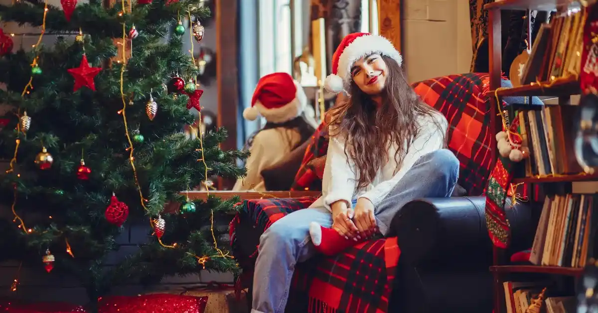 Young woman wearing a Santa hat, sitting on a cozy armchair with a red plaid blanket, smiling beside a decorated Christmas tree with ornaments and lights in a festive indoor setting