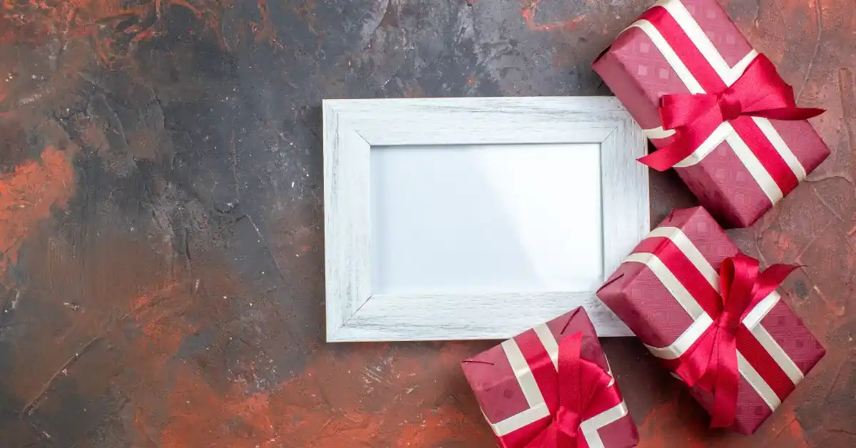 White blank photo frame placed on a textured brown background with three pink gift boxes wrapped in red ribbons arranged beside it