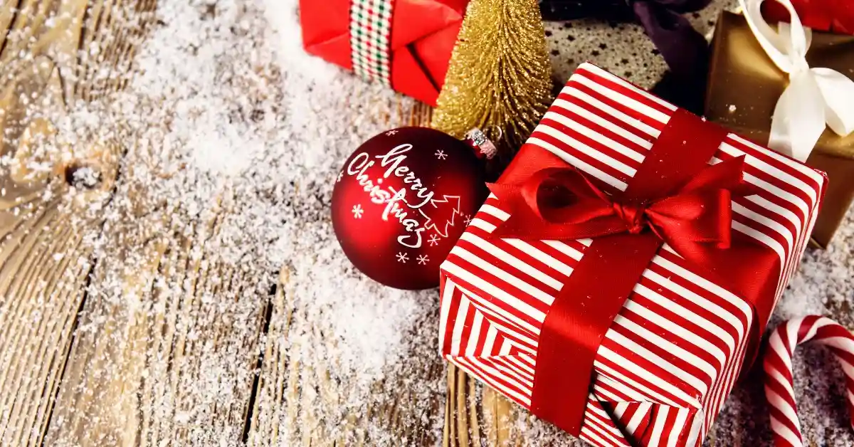 A red and white striped gift box with a red ribbon, Christmas decorations, and a red ornament on a snowy wooden surface