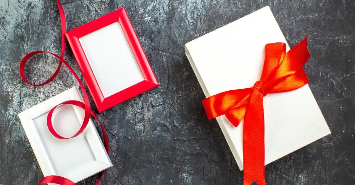 White gift box tied with a bright red ribbon placed on a dark textured background, with two empty photo frames and a loose red ribbon beside it.