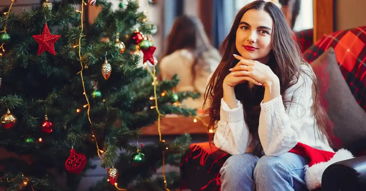 Young woman sitting beside a decorated Christmas tree with red ornaments and warm lights, smiling indoors in a cozy holiday setting