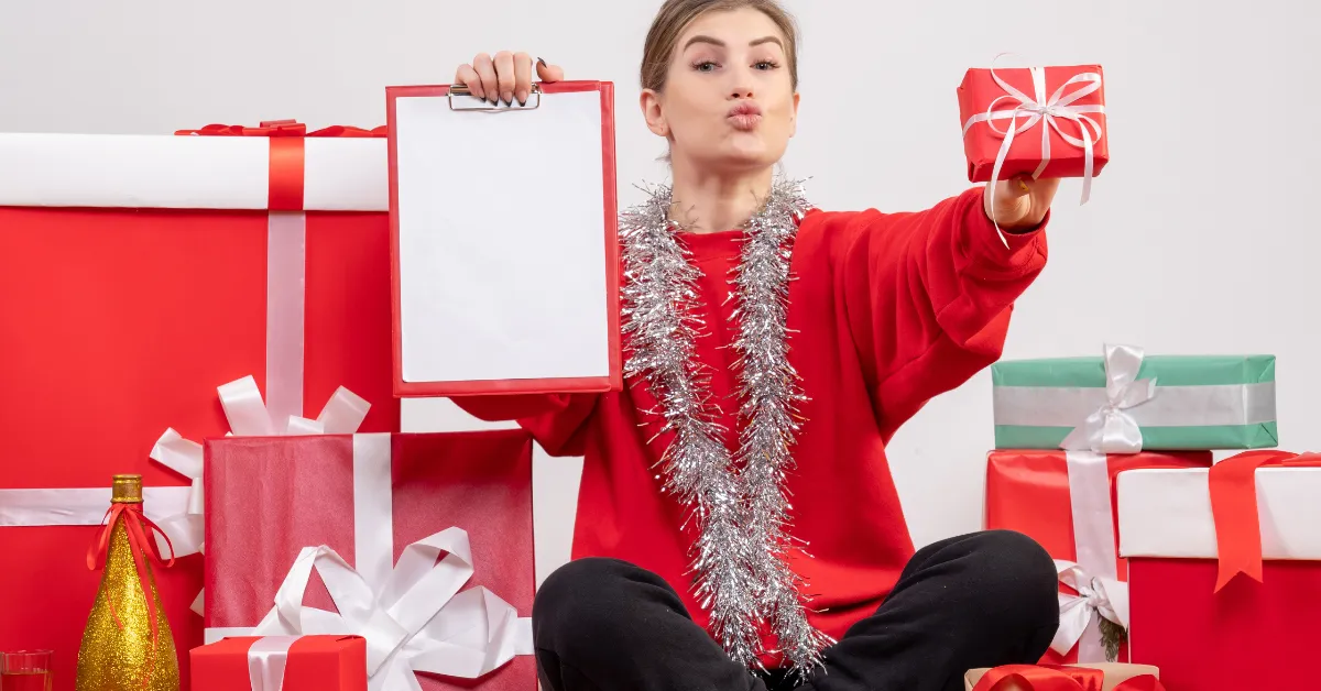 A woman wearing a red sweater and silver tinsel garland, sitting among wrapped Christmas gifts in red, white, and green. She holds a small gift in one hand and a blank clipboard in the other, making a playful kissing face