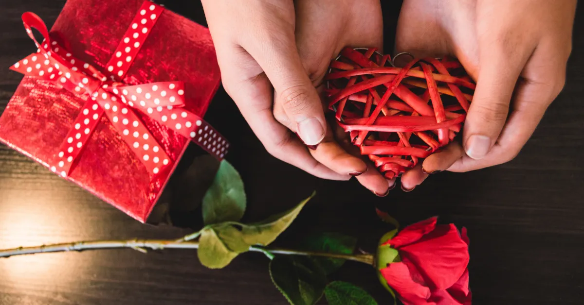Hands holding a red wicker heart, with a red gift box and a red rose on a dark wooden surface