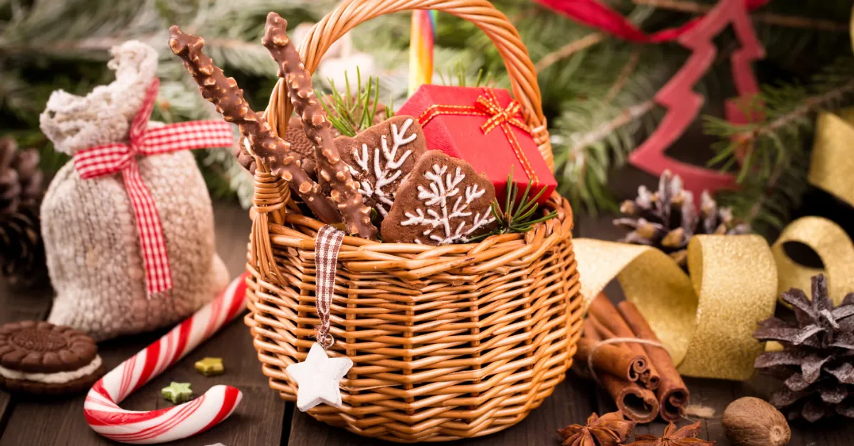 Christmas gift basket with gingerbread cookies, chocolate sticks, candy canes, and a small wrapped present surrounded by pine branches and holiday decorations.