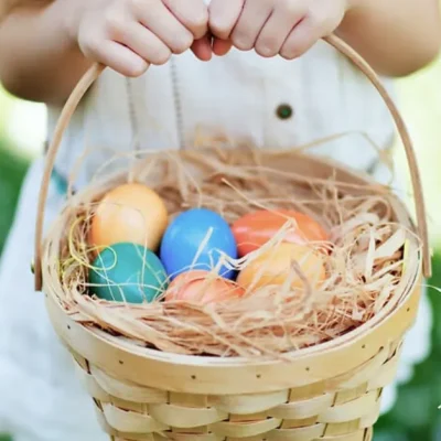 Child holding a wicker basket filled with colorful Easter eggs in grass during an outdoor Easter egg hunt.
