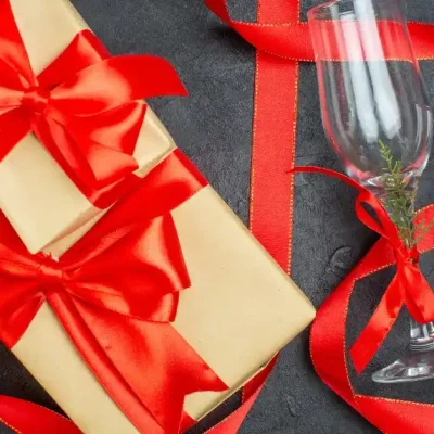 A gift box tied with a red ribbon and two champagne glasses, with red ribbons and small green sprigs on a dark background.