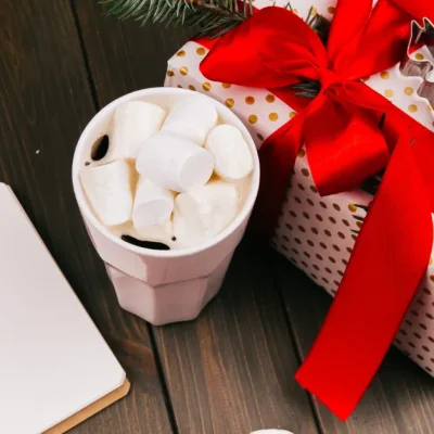Festive gift box with red ribbon placed on a wooden table beside a cup of hot chocolate with marshmallows and a blank notebook