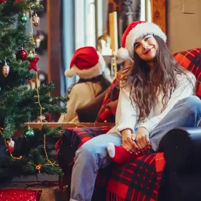 Young woman wearing a Santa hat, sitting on a cozy armchair with a red plaid blanket, smiling beside a decorated Christmas tree with ornaments and lights in a festive indoor setting