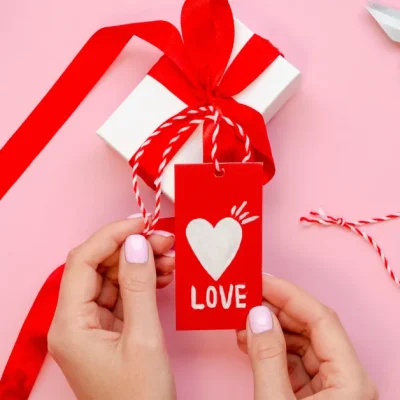 Hands holding a red ‘Love’ gift tag attached to a white present with red ribbon, surrounded by hearts, scissors, and wrapping accessories on a pink background.